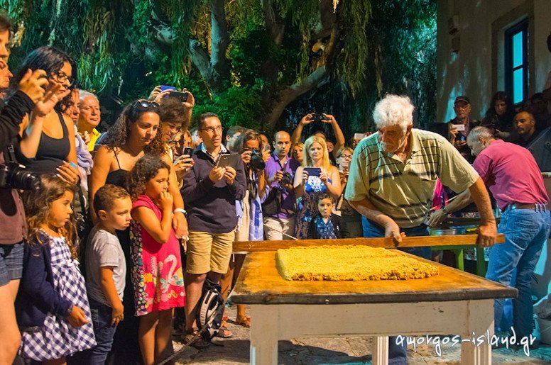 Amorgos, Chora, The Pasteli Festival - Gastronomy Tours man lying ‘pasteli’ composition on the table with a wooden spoon at the Pasteli Festival, Amorgos, Chora, Greece surrounded by people by night