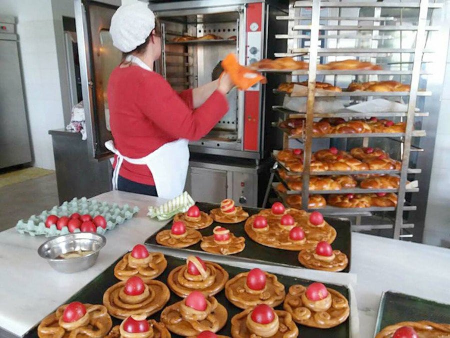 Avgoules - Gastronomy Tours Woman putting bakery pan with Greek Easter cakes on metal trolley and surrounded by sweets with red eggs on the top