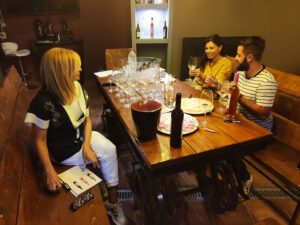 Three people stand in a dimly lit wine cellar, holding glasses and tasting different wines. The shelves are filled with bottles.