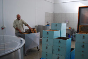 man placing honeycomb panel in honey extractor surrounded by bee hives and others apiculture machines at 'Ermionis'