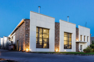 illuminated 'Katsaros Distillery' building by night with pavement and green lawn in the front